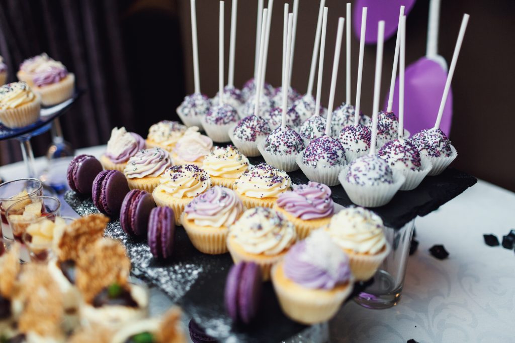 Elegantný candy bar s fialovými a bielymi cupcakes, fialovými makrónkami a bielymi cake pops zdobenými fialovým posypom, usporiadanými na čiernych bridlicových podnosoch.